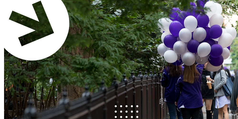 A student photographed from behind carrying purple and white balloons